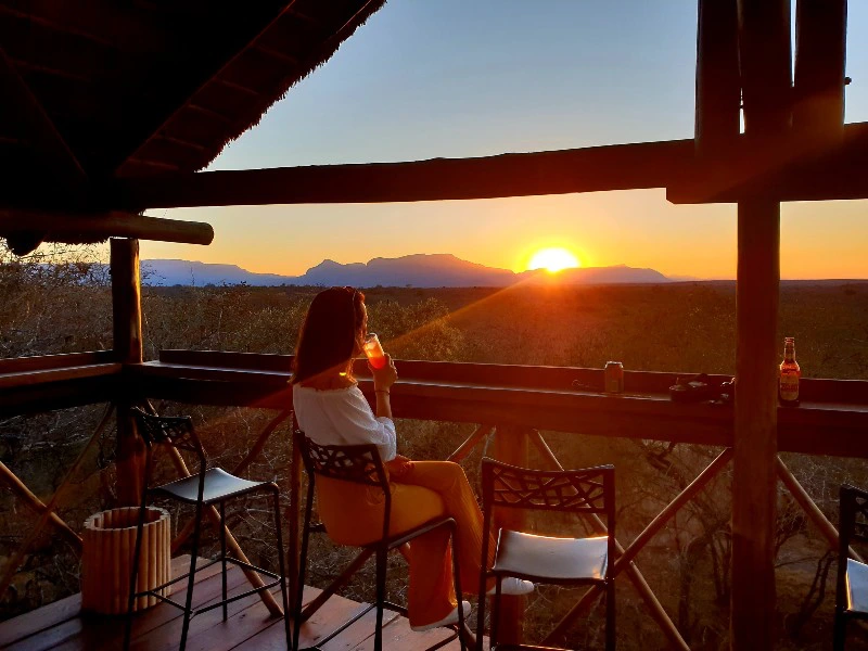 Vrouw zit op een terras met een drankje in de hand en kijkt naar de zonsondergang boven een heuvelachtig landschap.