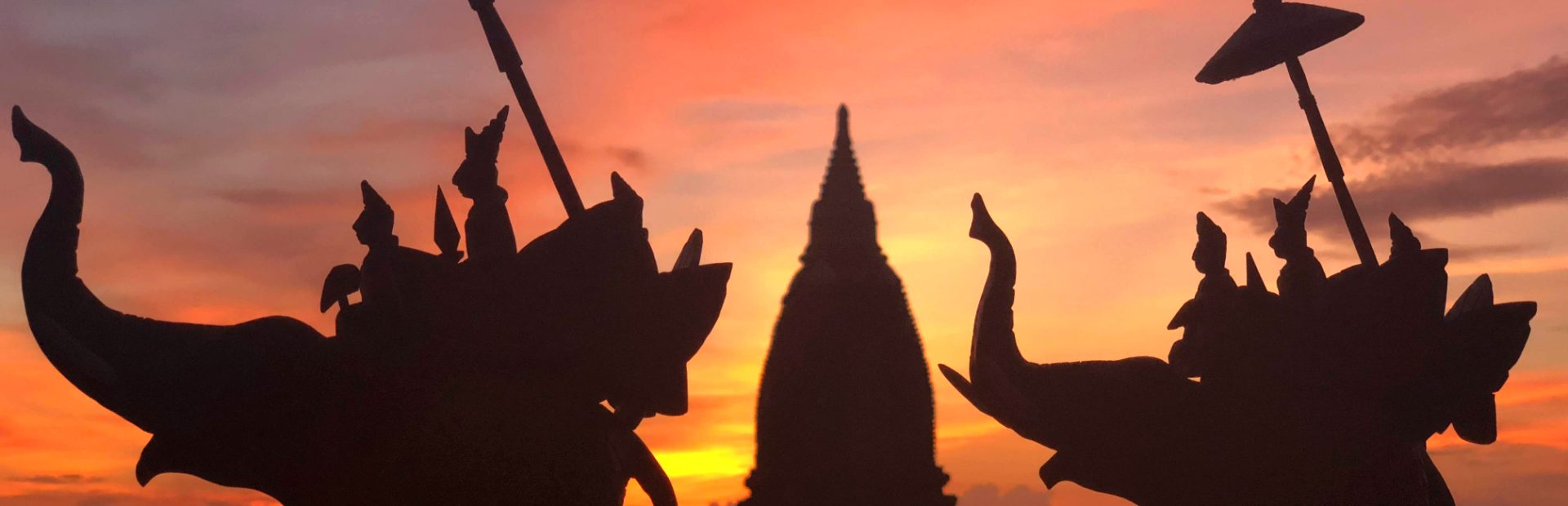 Silhouetten van ruiters op olifanten met speer en parasol bij zonsondergang voor een tempel.