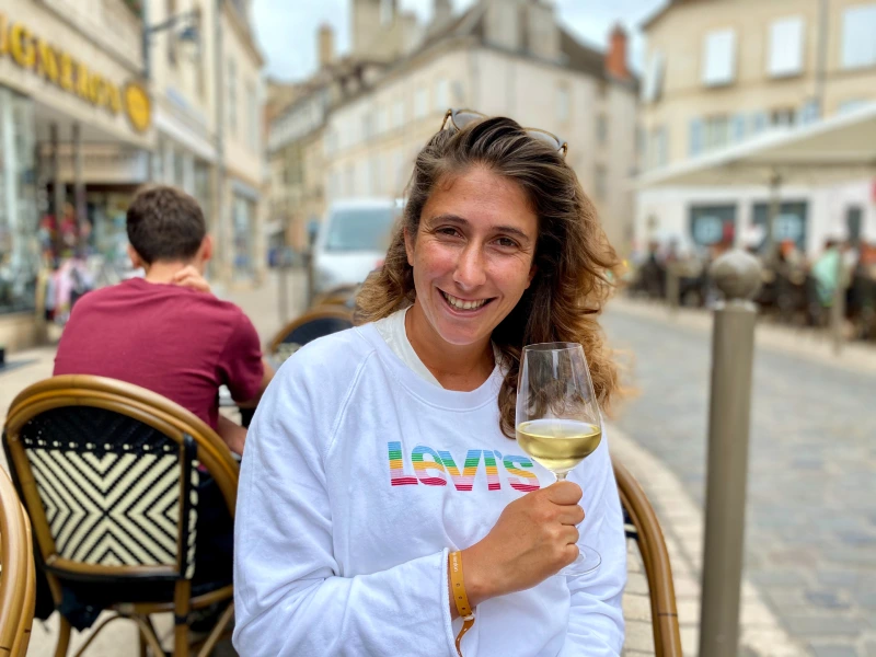 Vrouw zit op een terras in een Europese stad en houdt een glas witte wijn vast, met sfeervolle straat en terrashuisjes op de achtergrond.
