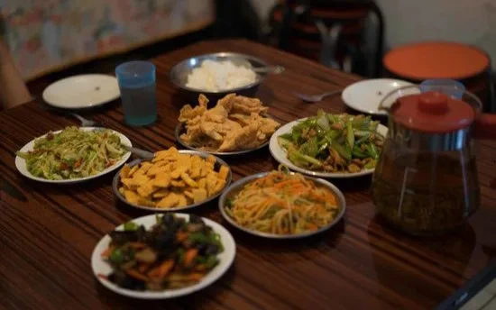 Tafel met verschillende schotels Chinees eten, waaronder groente, noedels, tofu en rijst, naast een theepot.