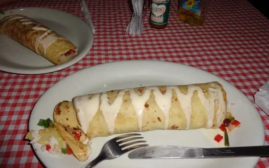 Grote burrito op een wit bord met saus eroverheen, geserveerd met mes en vork.