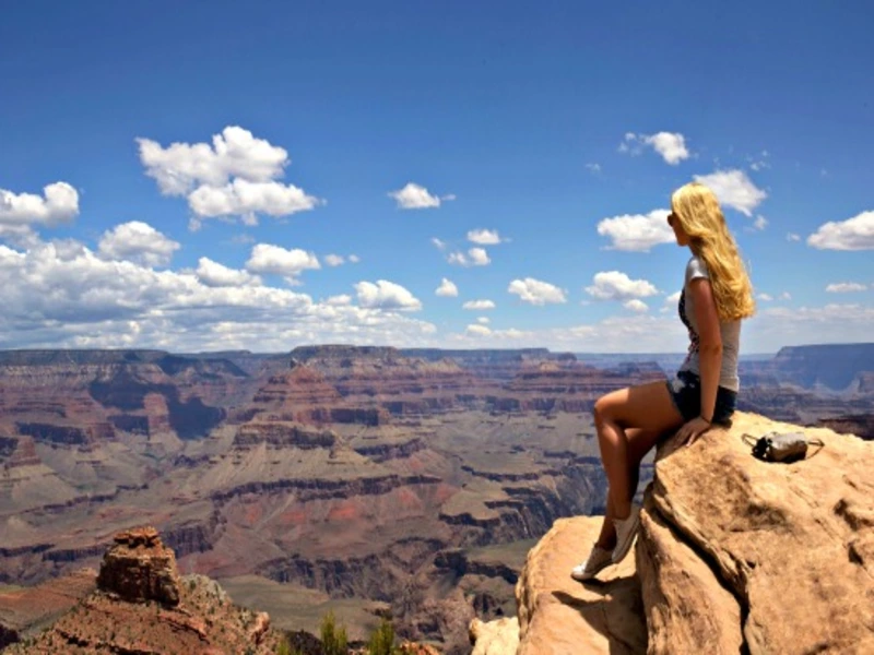 Vrouw zit op een rotspunt en kijkt uit over de uitgestrekte Grand Canyon onder een blauwe lucht met wolken.