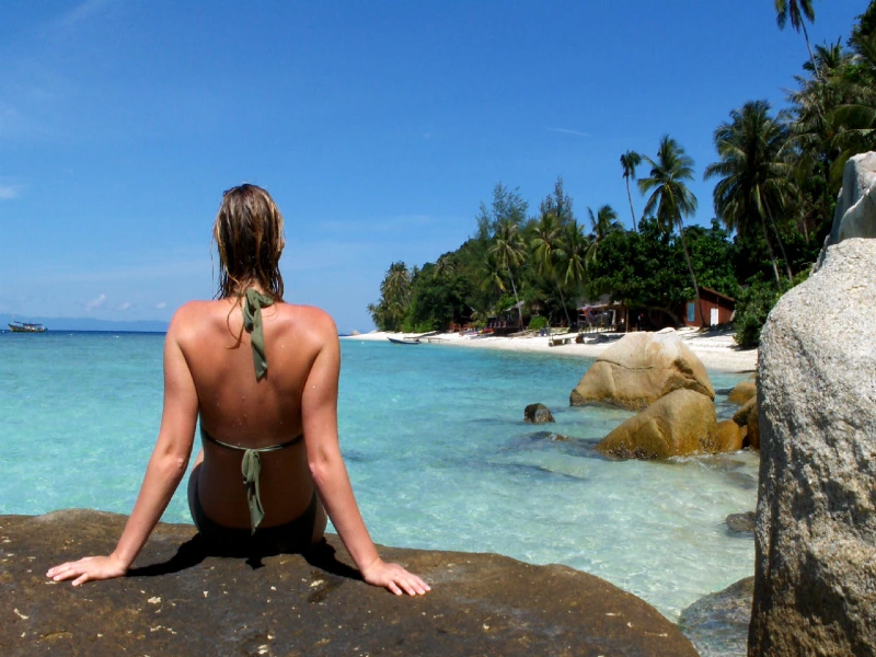 Vrouw in bikini zit op een rots en kijkt uit over turquoise water en een tropisch strand met palmbomen.