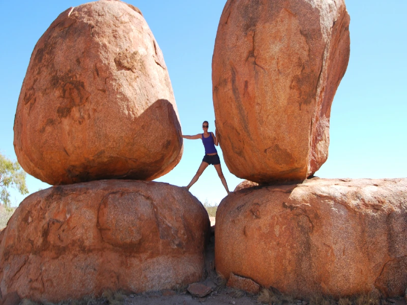 Reiziger poseert tussen twee enorme, ronde rotsblokken in de outback van Australië, bij de iconische Devil’s Marbles, onder een strakblauwe lucht.