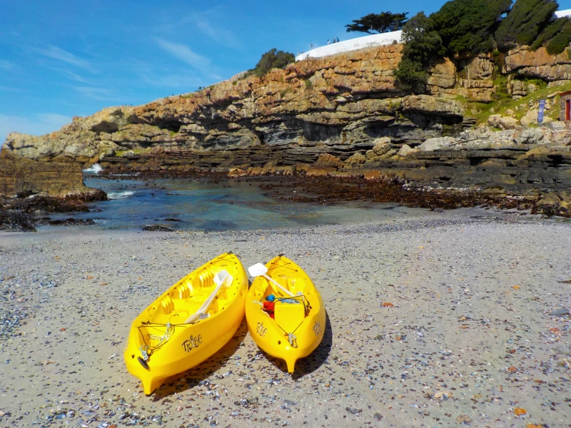 Twee gele kajaks liggen op een kiezelstrand met een rotsachtige kust en helderblauwe zee op de achtergrond.