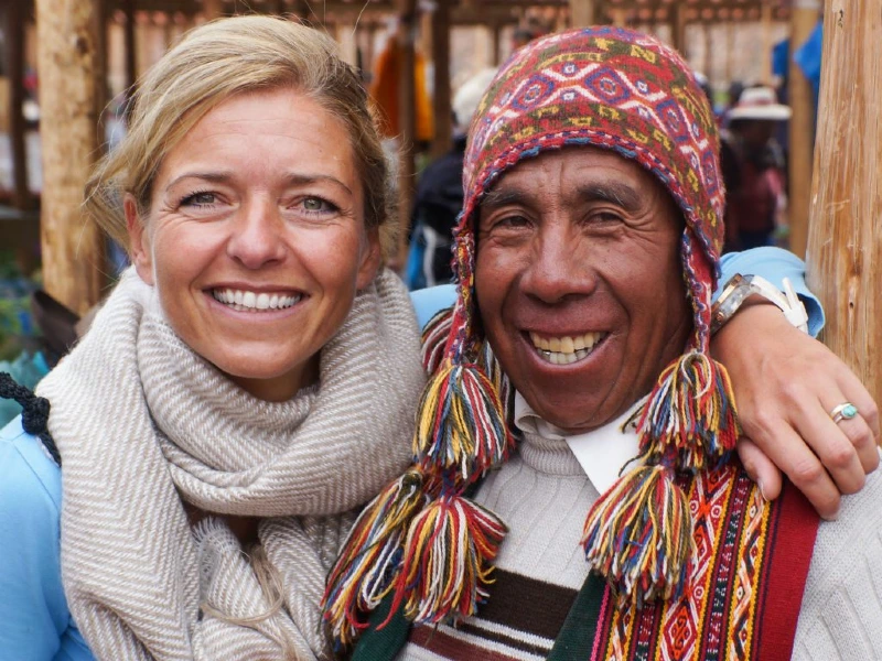 Twee lachende mensen schouder aan schouder: een reizigster en een lokale man in traditionele kleurrijke kleding en muts
