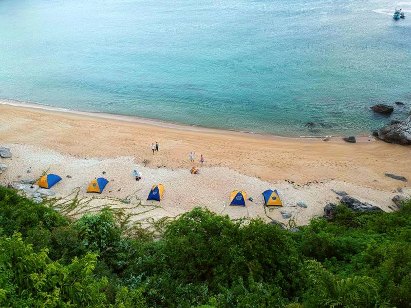 Rij met kleine tentjes opgesteld op een rustig strand aan zee, met enkele mensen die op het zand lopen.