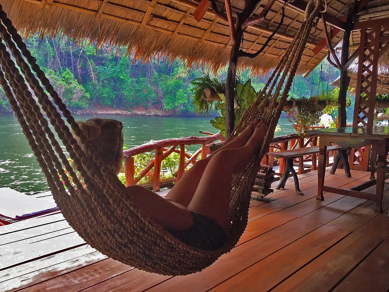 Persoon ligt in een hangmat onder een rieten dak op een houten veranda, met uitzicht op een rustige rivier en groene jungle op de achtergrond.