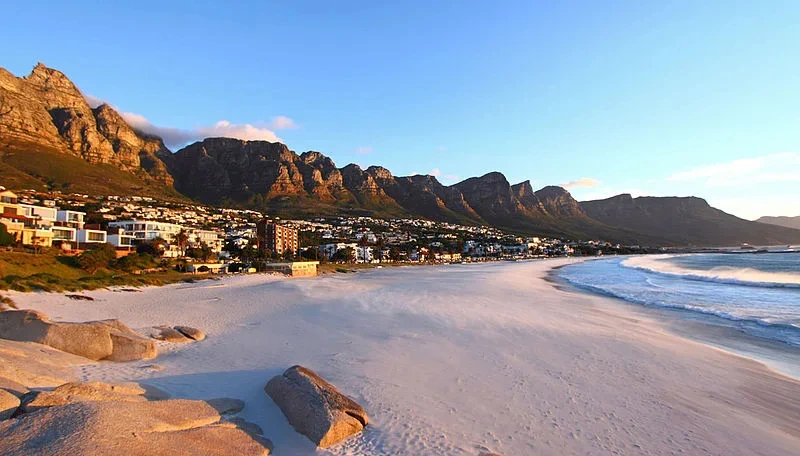 Uitzicht op Camps Bay in Kaapstad met een breed zandstrand, golven van de Atlantische Oceaan en de Twaalf Apostelen bergen op de achtergrond bij zonsondergang.