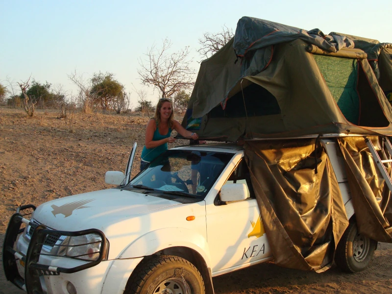 Vrouw klapt het daktentje uit bovenop een witte 4x4 met KEA-logo, geparkeerd in een droog, zanderig landschap met kale bomen