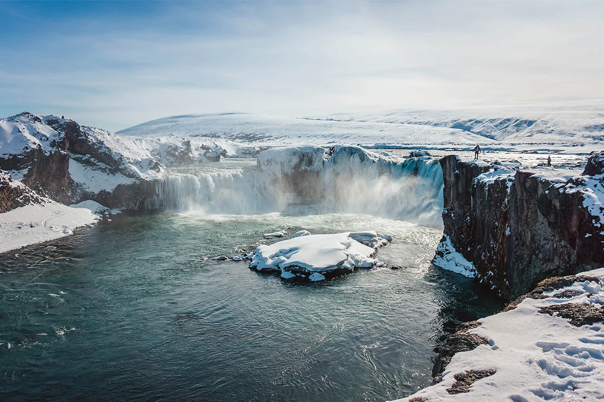 Bevroren waterval in IJsland, omgeven door besneeuwde rotsen en heuvels, met opspattend water en enkele mensen die op de klifrand staan.