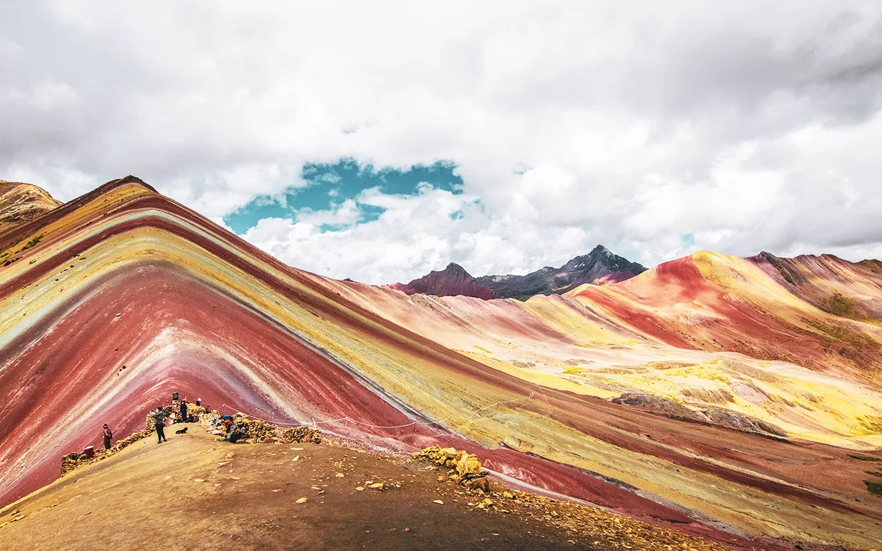 Kleurrijke Rainbow Mountain in Peru met lagen rood, geel en groen gesteente, omringd door wandelaars en een bergachtig landschap onder een deels bewolkte lucht.