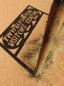 Schaduw van een bord met de tekst “Twyfelfontein Visitors Centre” op rood zand naast een houten paal.