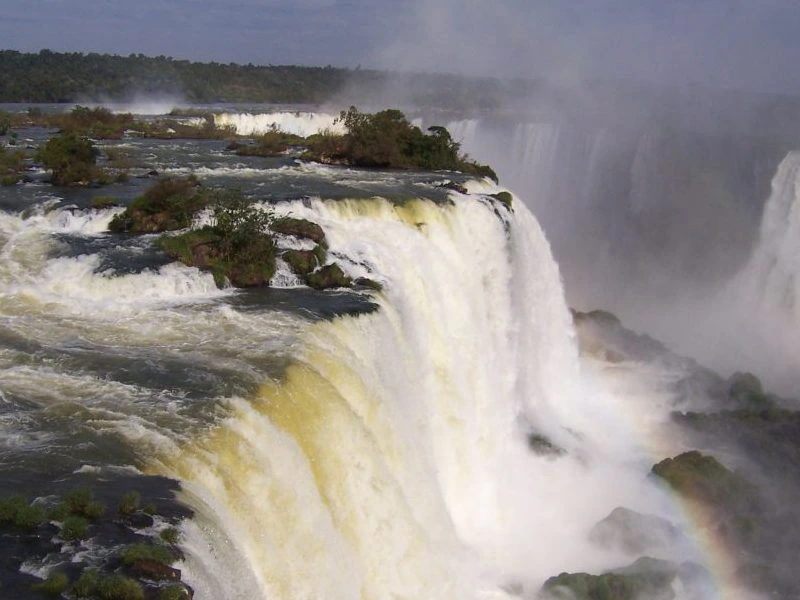 Grote watervallen storten naar beneden tussen rotsen, met mist en een regenboog langs de Iguazú-watervallen.