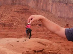Perspectiefgrap waarbij een man te paard in een rood rotslandschap lijkt te worden vastgehouden tussen duim en vingers van een hand op de voorgrond.