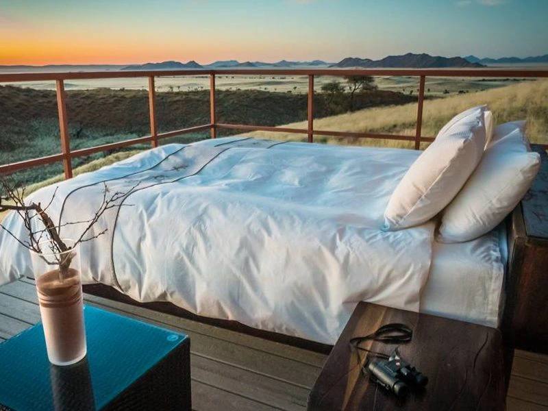 Buitenbed met wit beddengoed op een houten terras met uitzicht op een uitgestrekt landschap en zonsondergang, met een drankje en verrekijker op het tafeltje ernaast.