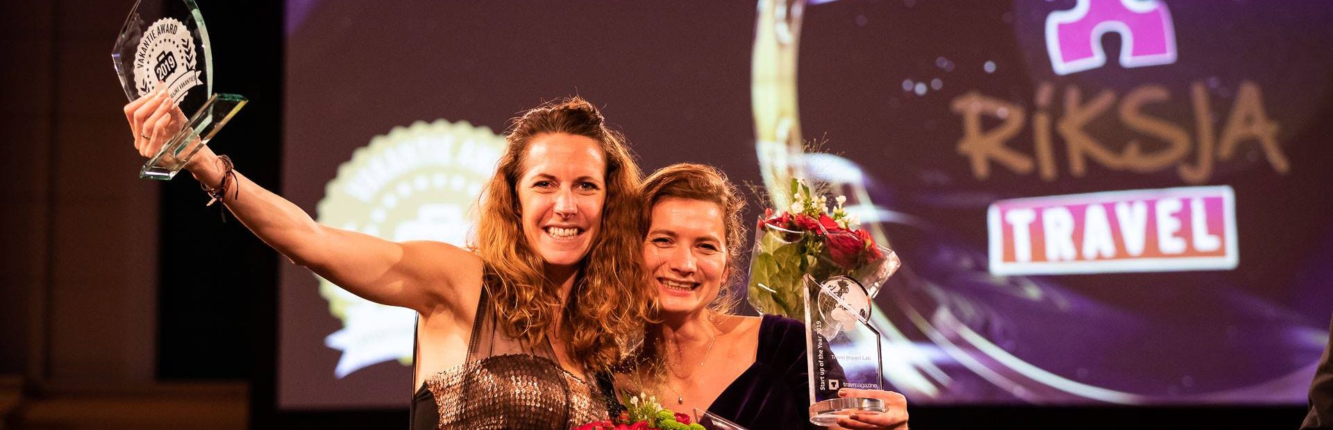 Twee vrouwen op een podium tijdens een prijsuitreiking, lachend met bloemen en glazen awards in hun handen, met op de achtergrond het Riksja Travel-logo geprojecteerd.
