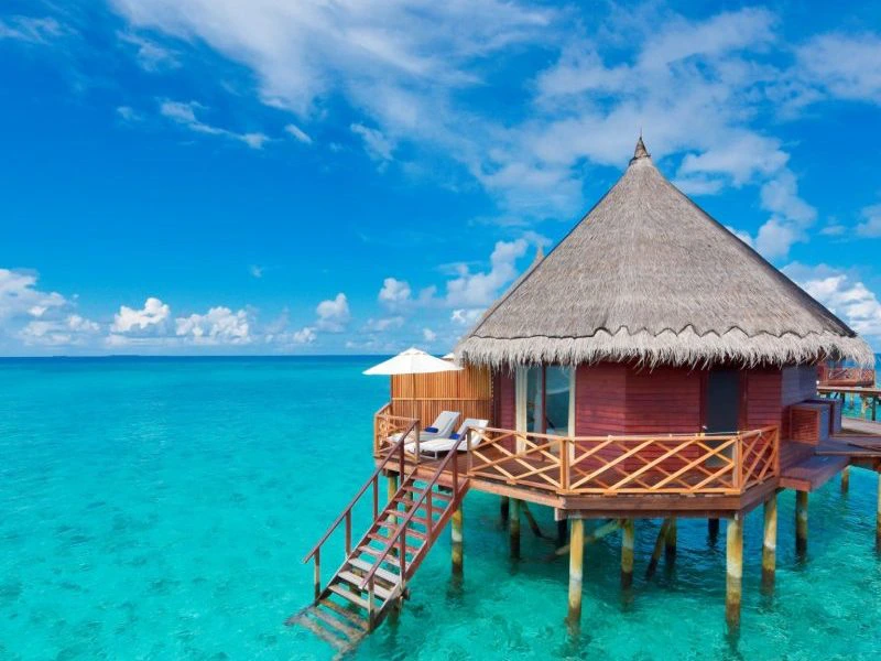 Een luxe waterbungalow met rieten dak op palen in helderblauw water, met een houten terras en ligstoelen, onder een zonnige blauwe lucht.