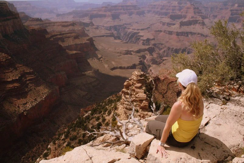 Vrouw met gele top en witte pet zit op de rand van een rots en kijkt uit over de Grand Canyon.