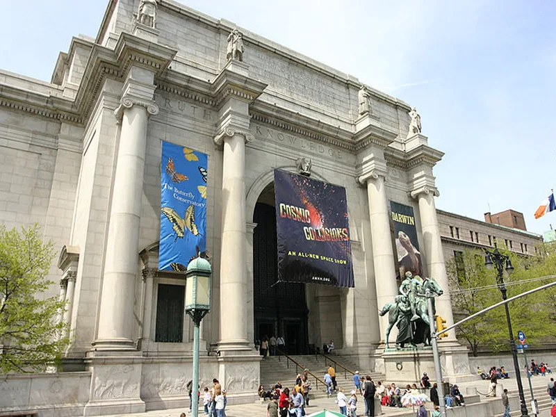 Voorkant van het American Museum of Natural History in New York, met hoge pilaren, banners van tentoonstellingen en mensen op de trappen.