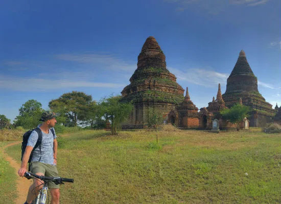 Persoon met een fiets kijkt naar oude boeddhistische tempels in Bagan, Myanmar, omgeven door gras en bomen onder een heldere blauwe lucht.