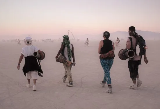 Vier mensen lopen over een stoffige vlakte bij zonsondergang met muziekinstrumenten op de rug.