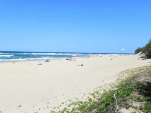 Uitgestrekt zandstrand met mensen die verspreid liggen te zonnen en zwemmen in zee, onder een strakblauwe lucht.