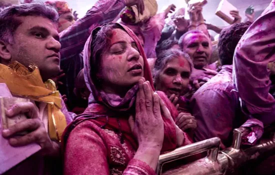 Mensen bedekt met felgekleurde poeder tijdens het Holi-festival, met een vrouw op de voorgrond die met gesloten ogen en gevouwen handen bidt.