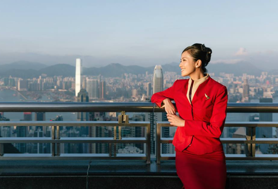 stewardess in rood uniform leunt ontspannen tegen een reling met uitzicht op de skyline van Hongkong