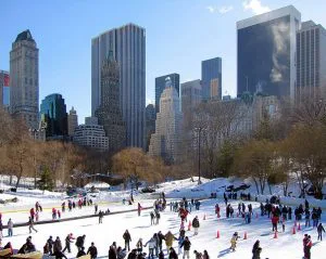 Schaatsers genieten van de ijsbaan in Central Park, New York, met besneeuwde bomen op de voorgrond en hoge wolkenkrabbers op de achtergrond.