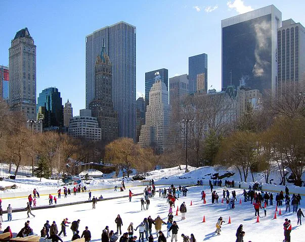 Schaatsers genieten van de ijsbaan in Central Park, New York, met besneeuwde bomen op de voorgrond en hoge wolkenkrabbers op de achtergrond.