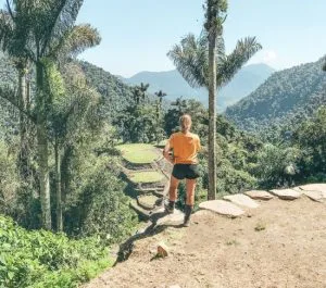 Vrouw kijkt uit over de groene terrassen van de verloren stad Ciudad Perdida in de jungle van Colombia, omringd door palmbomen en bergen.
