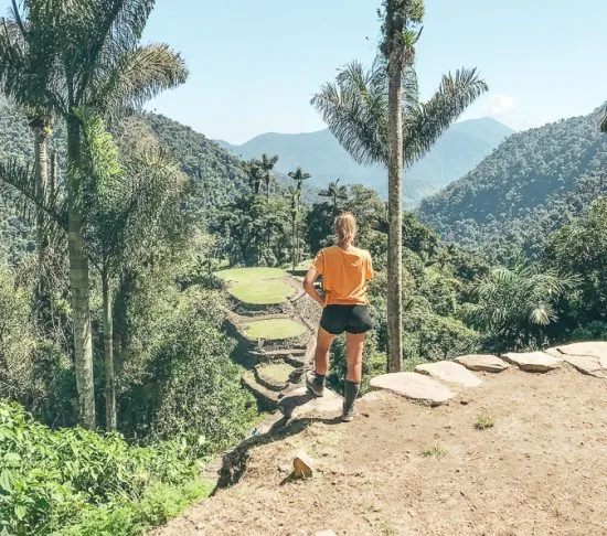 Vrouw kijkt uit over de groene terrassen van de verloren stad Ciudad Perdida in de jungle van Colombia, omringd door palmbomen en bergen.