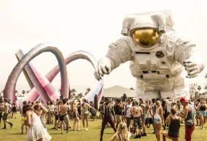 Festivalterrein met dansende mensen, een groot astronautenbeeld en kleurrijke kunstinstallatie.