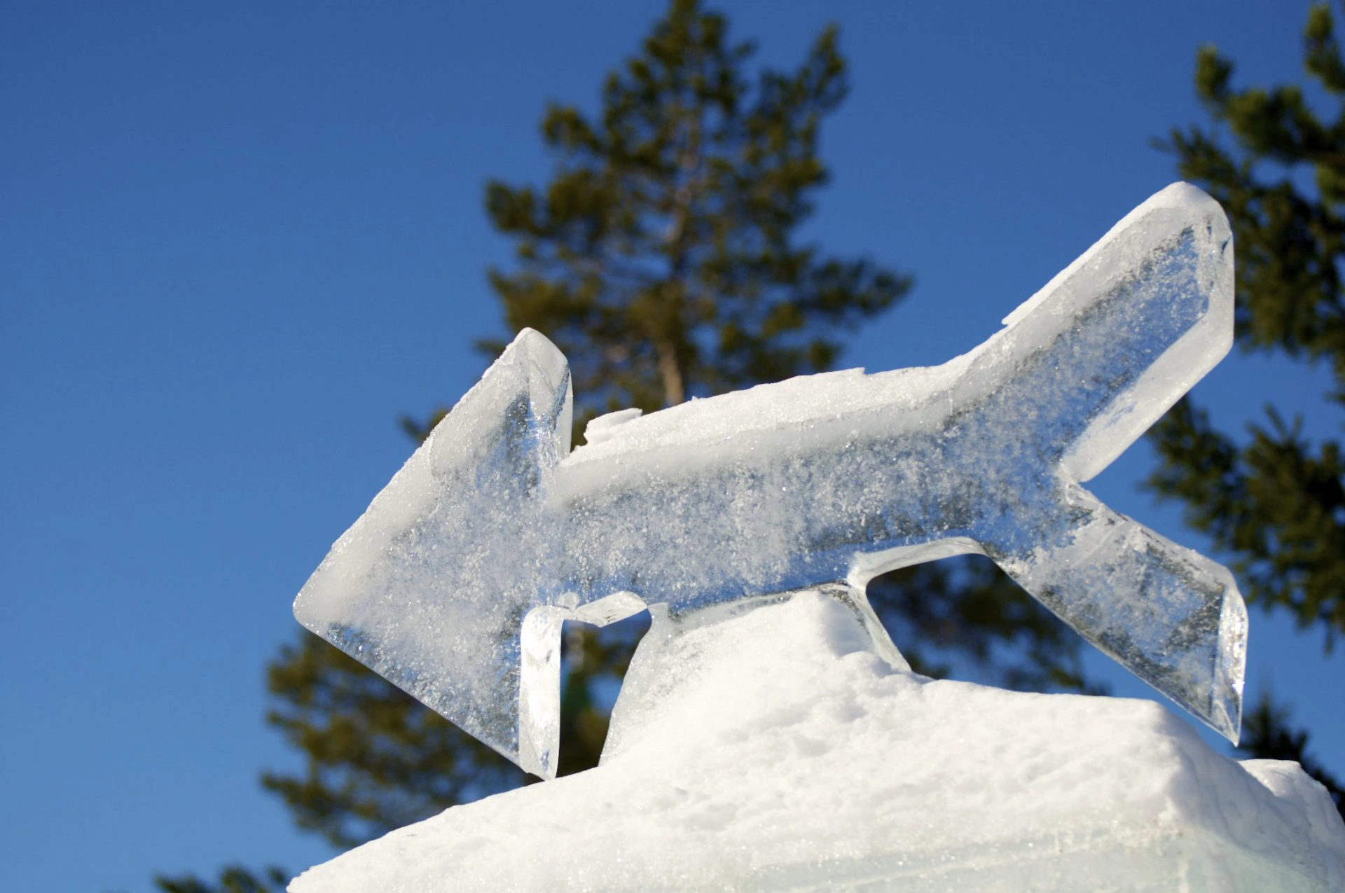 IJssculptuur in de vorm van een pijl die naar links wijst, glinsterend in de zon met een blauwe lucht en bomen op de achtergrond.