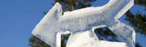 IJssculptuur in de vorm van een pijl die naar links wijst, glinsterend in de zon met een blauwe lucht en bomen op de achtergrond.