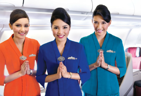 Drie lachende stewardessen van Thai Airways in kleurrijke uniformen (oranje, paars en turquoise) begroeten de passagiers met gevouwen handen aan boord van het vliegtuig