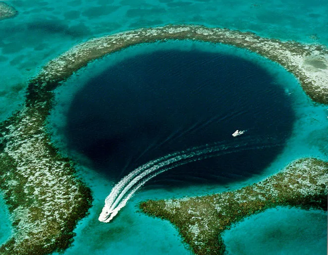 Luchtfoto van de Great Blue Hole in Belize, een grote ronde donkerblauwe onderwatergrot omringd door turquoise water en koraal, met twee boten die eroverheen varen.