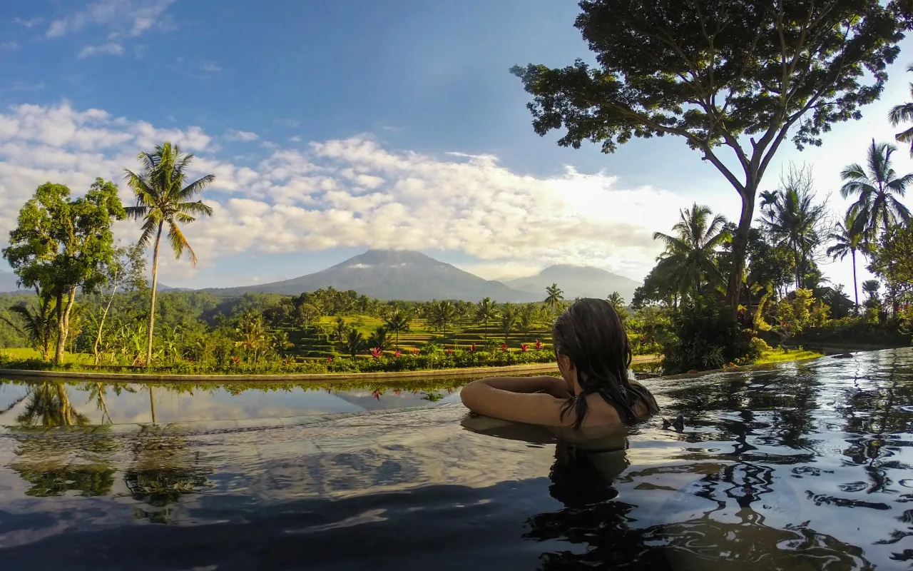 Vrouw ontspant in een infinity pool met uitzicht op een groene vallei en een vulkaan in de verte, omringd door palmbomen en tropische natuur.