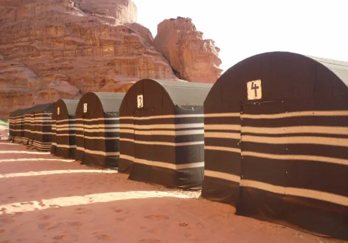 Rijtje traditionele zwarte woestijntenten met witte strepen in een zandige omgeving, tegen de achtergrond van rode rotsen in Jordanië.
