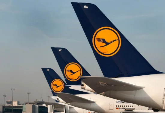 Staartstukken van Lufthansa-vliegtuigen op een rij, met het kenmerkende gele logo van een kraanvogel op een donkerblauwe achtergrond, geparkeerd op het vliegveld.