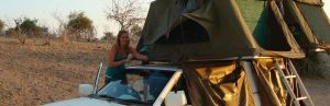 Vrouw zet een daktent op een witte jeep op in een droog, savanneachtig landschap.