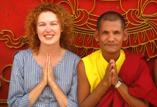 Een vrouw en een boeddhistische monnik zitten naast elkaar voor een rood versierd decor, beide met gevouwen handen in een begroetingsgebaar en een glimlach.