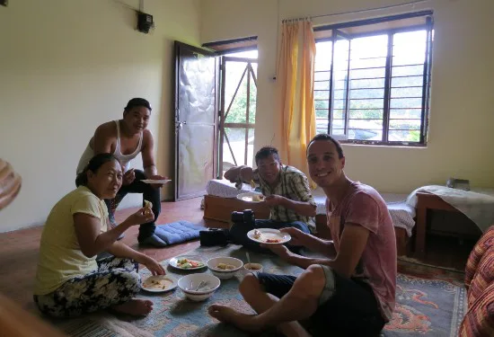 Vier mensen zitten op de grond in een woonkamer rond een kleed en eten samen van borden met traditioneel Nepalees eten, terwijl ze naar de camera glimlachen.