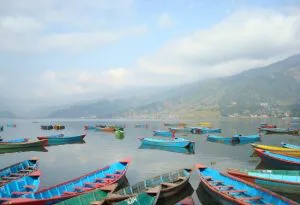Kleurrijke houten bootjes drijven op een rustig meer in Nepal, met groene heuvels en bergen op de achtergrond onder een deels bewolkte lucht.