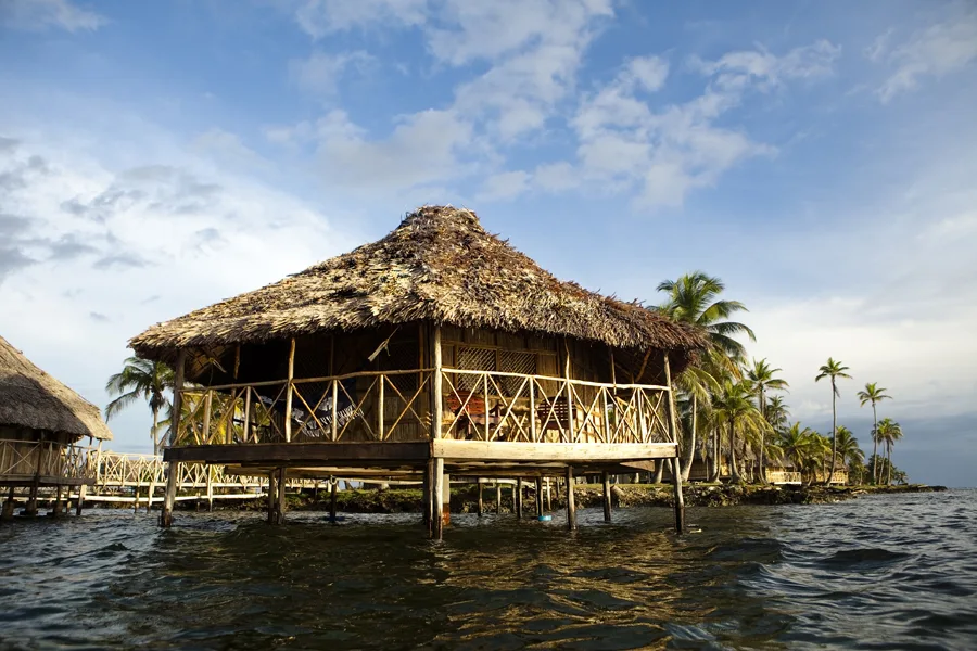 Traditioneel houten huisje op palen boven het water, omringd door palmbomen en een rustige zee, met een heldere lucht op de achtergrond.