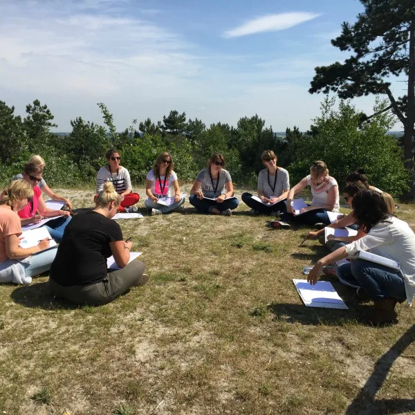 Groep mensen zit in een kring op een open plek in de natuur, schrijvend in notitieblokken tijdens een buitensessie.