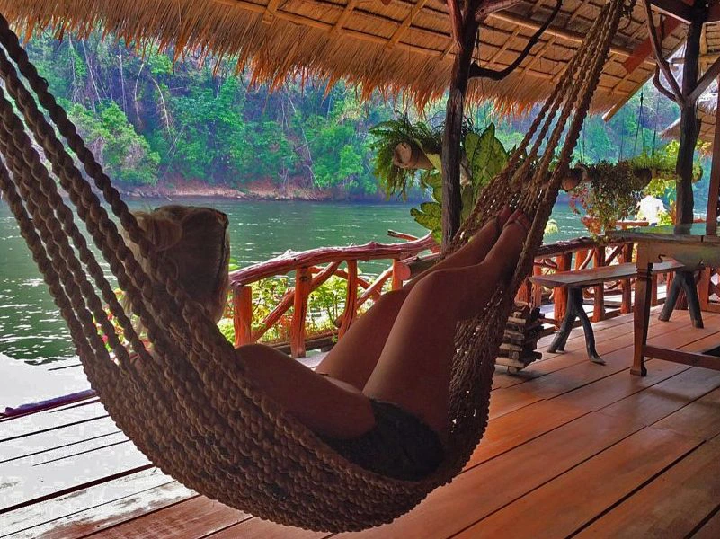 Persoon ligt in een hangmat onder een rieten dak op een houten veranda, met uitzicht op een rustige rivier en groene jungle op de achtergrond.