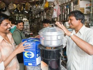 Mannen in een Indiase winkel demonstreren een energiezuinig kooktoestel met een aluminium pan erop, omringd door keukengerei en verpakkingen