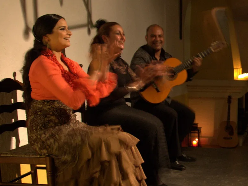 Drie artiesten voeren een traditionele flamenco-voorstelling op, met een gitarist en twee vrouwen die in kleurrijke jurken zingen en ritmisch klappen.